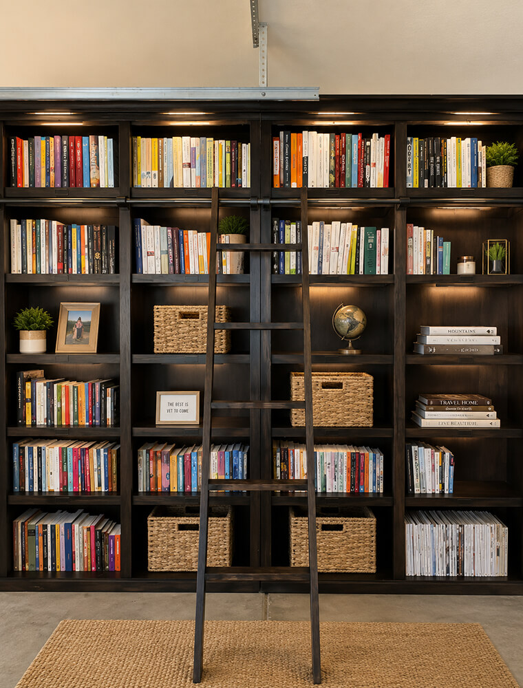 Floor-to-ceiling dark wood home library organized by Lifelystyle in Las Vegas — built-in lighting, rolling library ladder, woven baskets, and curated book arrangements that turn a bookcase into a statement